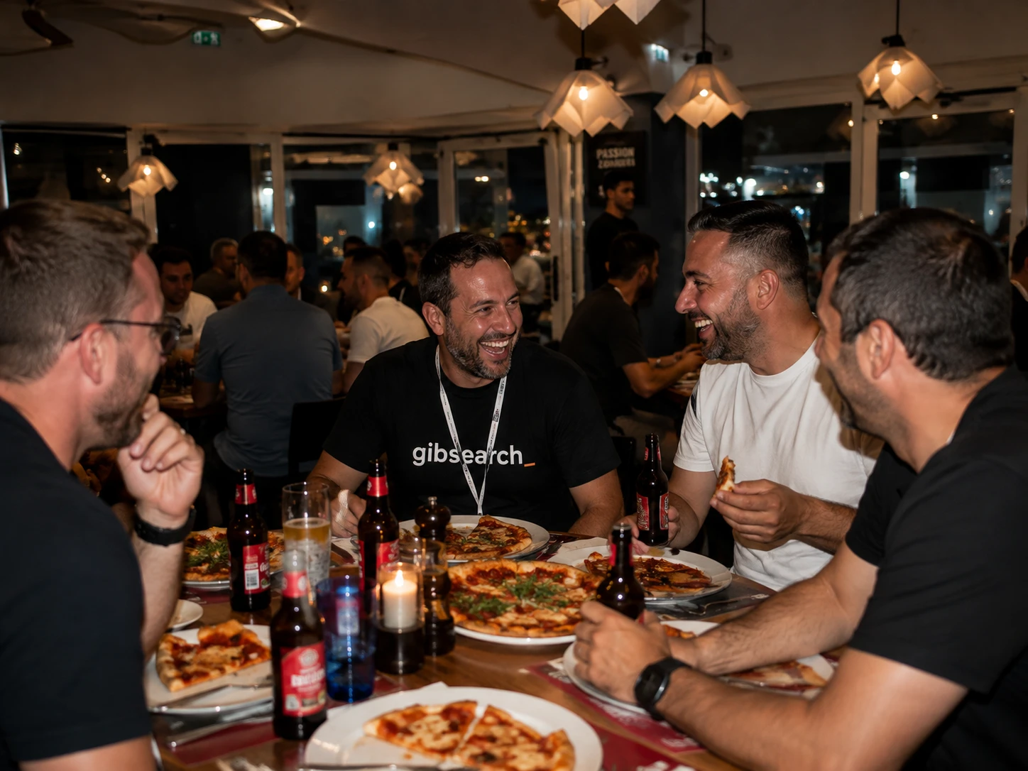 A group laughing over food and beers at a packed GibSearch table.